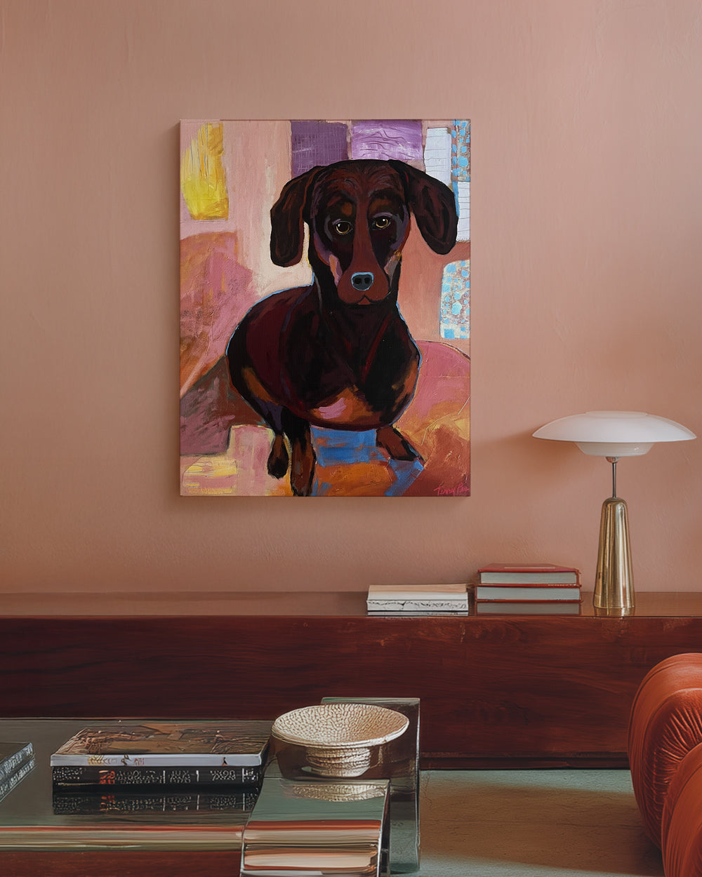 Painting of a dachshund on a wall above a wooden console table with books and a lamp. "Bella" is an original mixed media painting that captures the warmth and personality of a Dachshund in expressive, painterly detail. Layers of rose, amber, and deep brown create depth and character, while soft highlights bring the dogÕs soulful eyes to life. The mix of color and texture adds a contemporary edge to a classic portrait, making this a standout piece for dog lovers and modern art collectors alike.